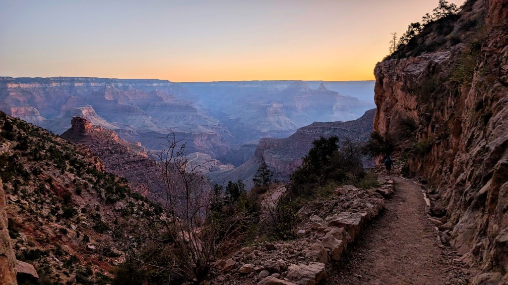 Bright Angel Trail at sunrise: the path cuts along a cliff edge above layered Grand Canyon walls, warm orange light on the horizon and a lone hiker ahead on the trail