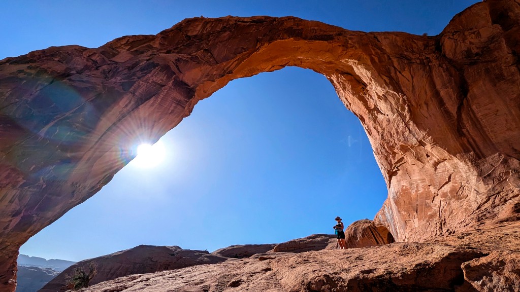 Massive orange sandstone Corona Arch near Moab, Utah, with a sunburst behind the arch and a hiker below for scale under a clear blue sky