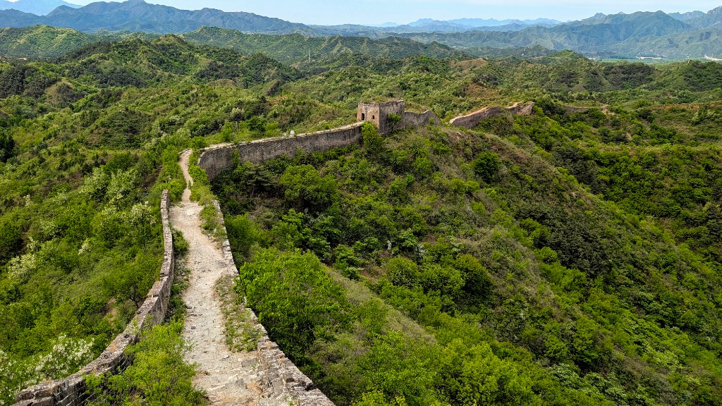 Unrestored Great Wall at Gubeikou – crumbling stone battlements overgrown with grass along a forested ridge