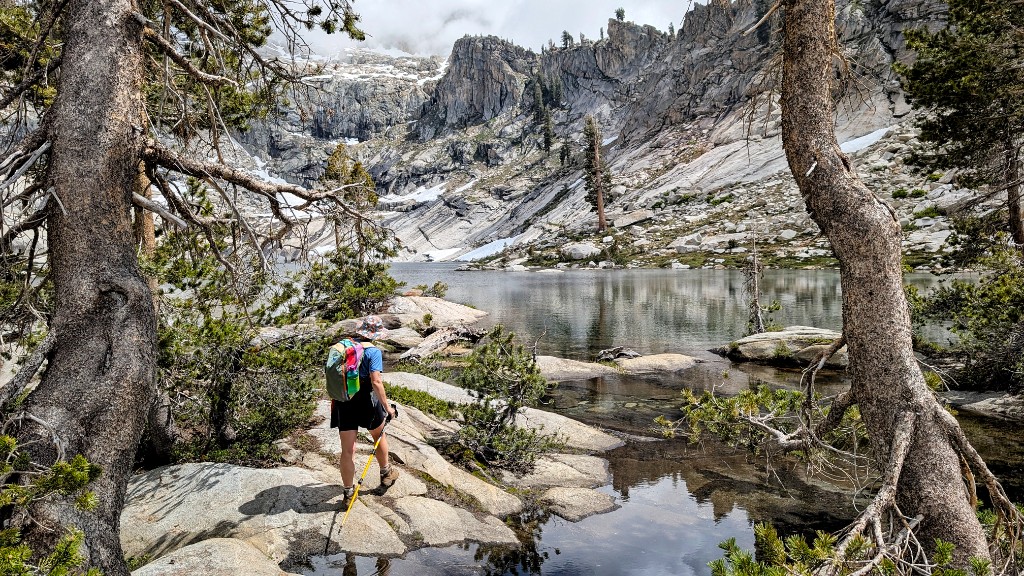 Hiker stepping across granite slabs beside Pear Lake in Sequoia National Park, with alpine cliffs and lingering snow in the background